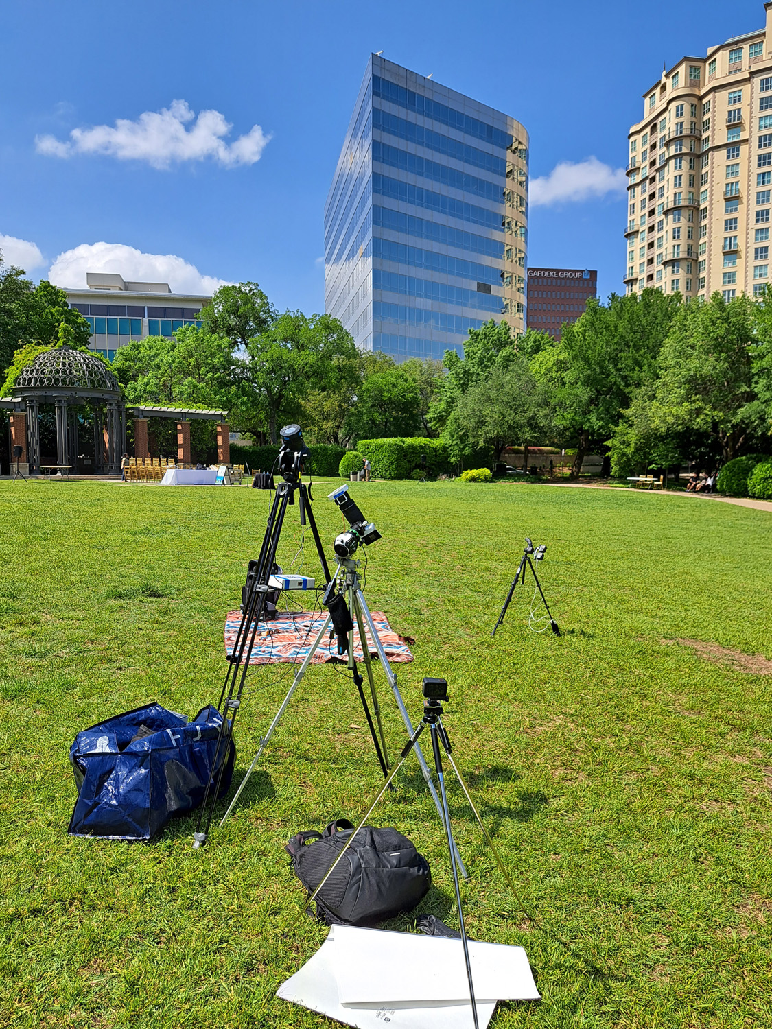 Multiple tripods and cameras on an empty green field, surrounded by trees and buildings.