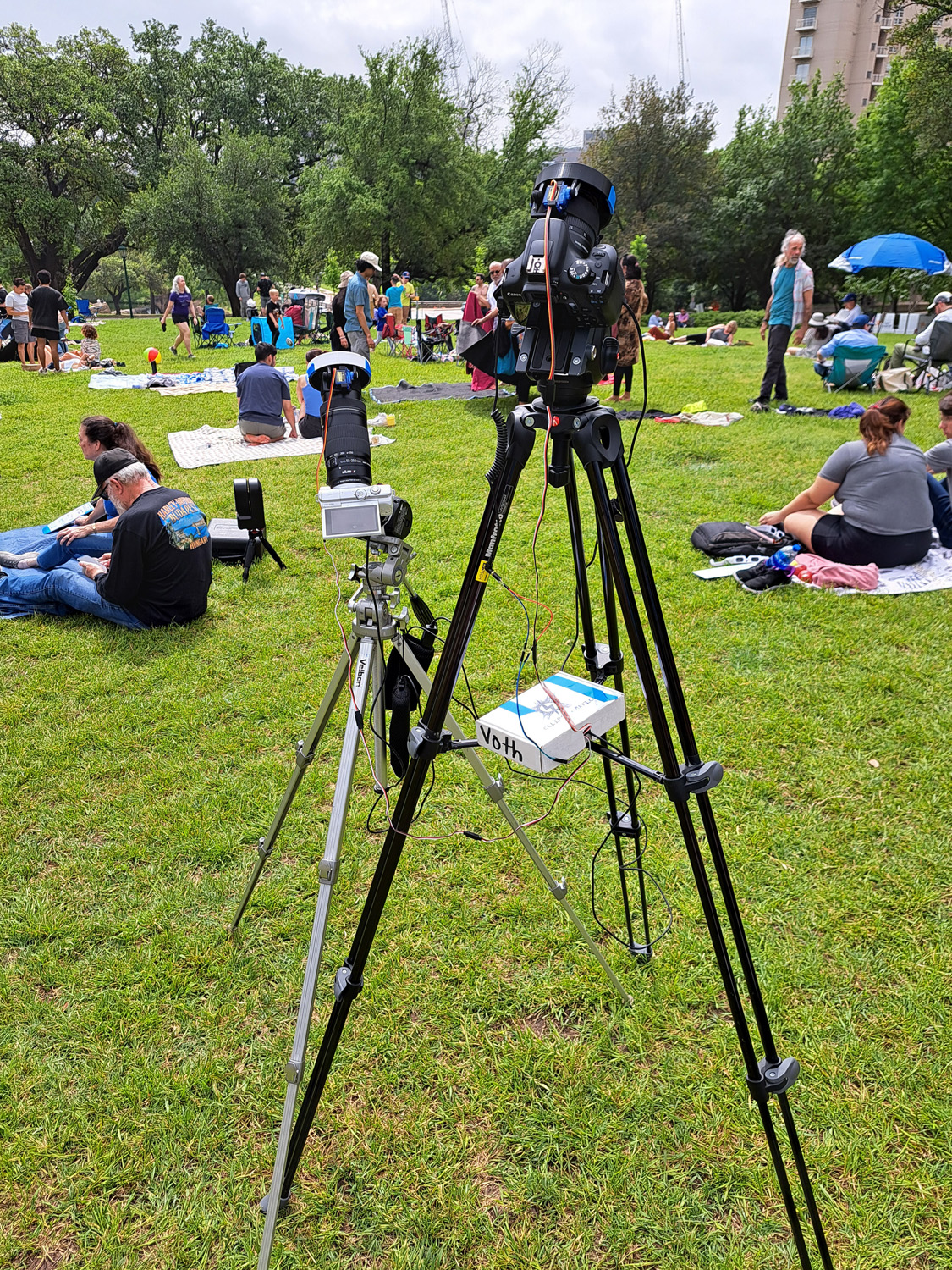 Two tripods and cameras, each with 3D printed solar filters and cables streaming into a white cardboard box. Grassy field with people in background.