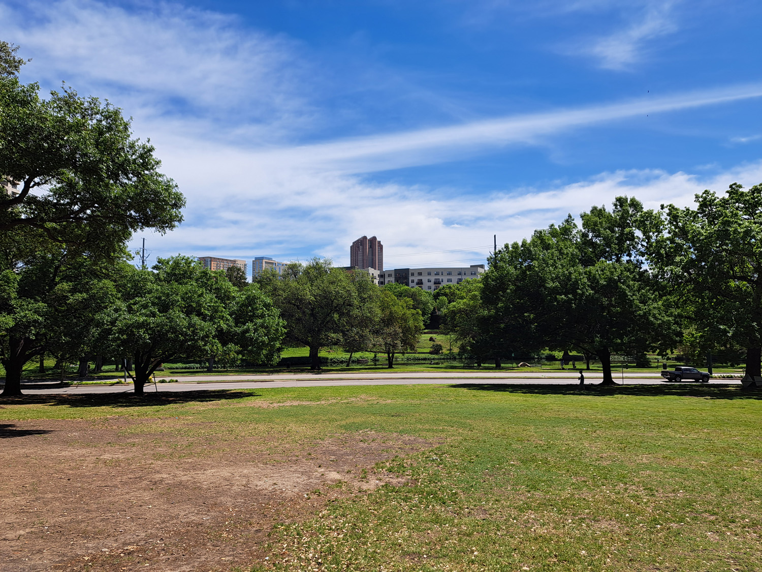 A grassy field enclosed by trees with a river and building in the background, under mostly clear skies.